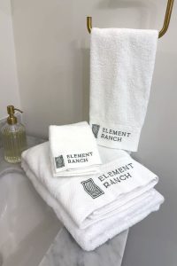 The image shows a set of white towels embroidered with the text "Element Ranch." They are neatly arranged in a bathroom setting, with one towel hanging on a gold-colored rail and others stacked on a marble countertop beside a soap dispenser. White towels labeled "Element Ranch" arranged in bathroom with marble counter and gold rail.