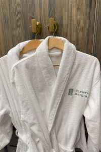 Two white bathrobes with "Element Ranch" embroidery are hanging on wooden hangers against a wood-textured wall. Two white bathrobes on hangers, embroidered "Element Ranch," against a wood-textured wall.