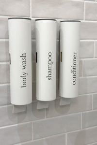Wall-mounted dispensers labeled for body wash, shampoo, and conditioner are seen against a tiled background. Wall-mounted dispensers for body wash, shampoo, and conditioner on a tiled wall.