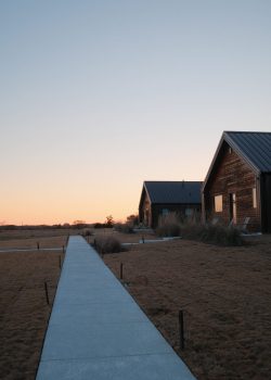 The image depicts a tranquil scene with a concrete path leading towards two wooden cabins under a clear sky at sunset. The landscape surrounding the path is grassy, with some shrubs near the cabins. Concrete path leading to two wooden cabins under clear sunset sky, surrounded by grass and shrubs.