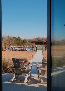 The image shows a view through a window of an outdoor patio area with two wooden chairs facing a walkway. The walkway leads to a fenced area with a pergola and a pool. The surrounding area features natural grasses and trees under a clear blue sky. View of outdoor patio with two wooden chairs, walkway to pergola, pool, grasses, and trees.