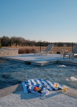 A poolside scene featuring a couple of lounge chairs on a deck overlooking a swimming pool. In the foreground, a blue and white striped towel, a pair of sunglasses, and two glasses with drinks are placed near the water. The background shows a fenced-off area with trees and grass under a clear sky. Poolside with chairs, towel, sunglasses, drinks, fenced area, trees, and clear sky.