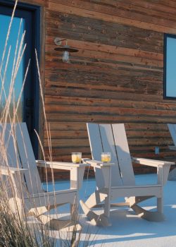 A wooden exterior of a building features a small porch area with two beige Adirondack chairs. The chairs have glasses with drinks placed on the armrests. Tall grasses are in the foreground, adding a natural element to the outdoor scene. Wooden building exterior with porch, two beige Adirondack chairs, drinks, and tall grasses.